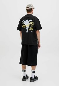 Zwart oversized t-shirt met palmboom grafiek en tekst op de achterkant, gecombineerd met zwarte shorts tot de knie en zwarte sneakers.