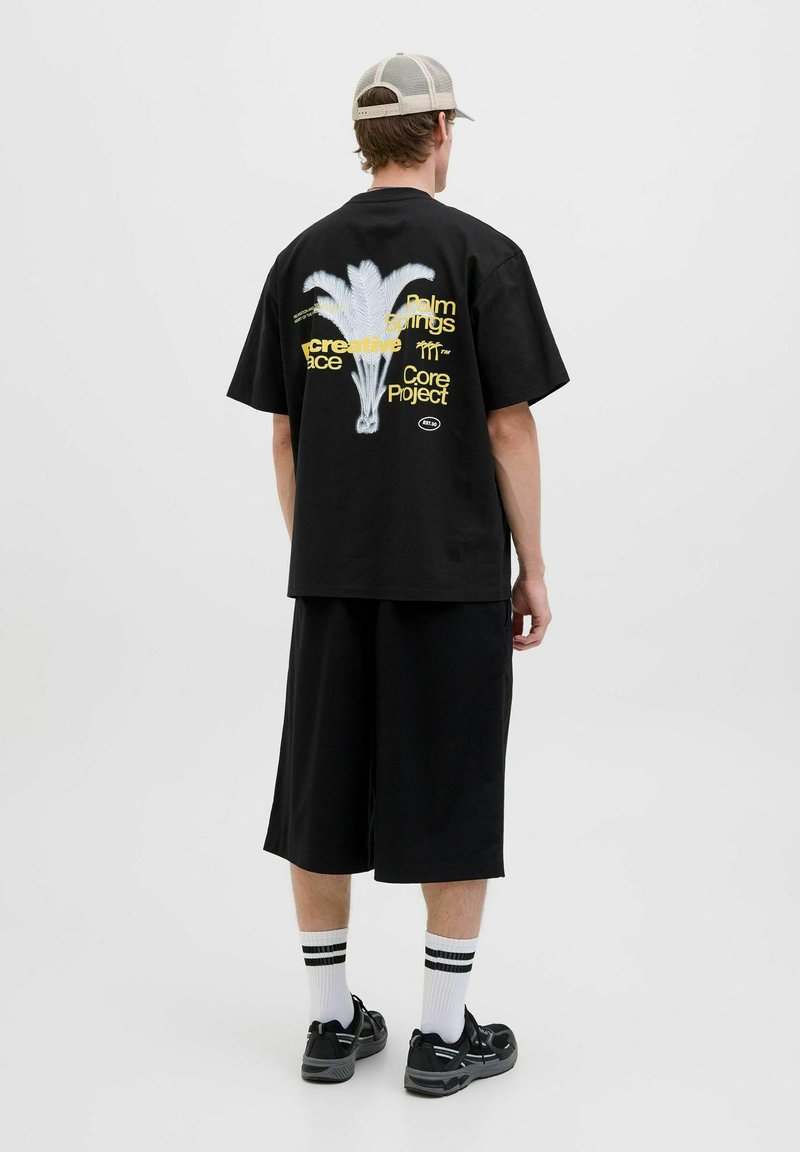 Sort oversized t-shirt med palmegrafik og tekst på bagsiden, parret med sorte knælange shorts og sorte sneakers.