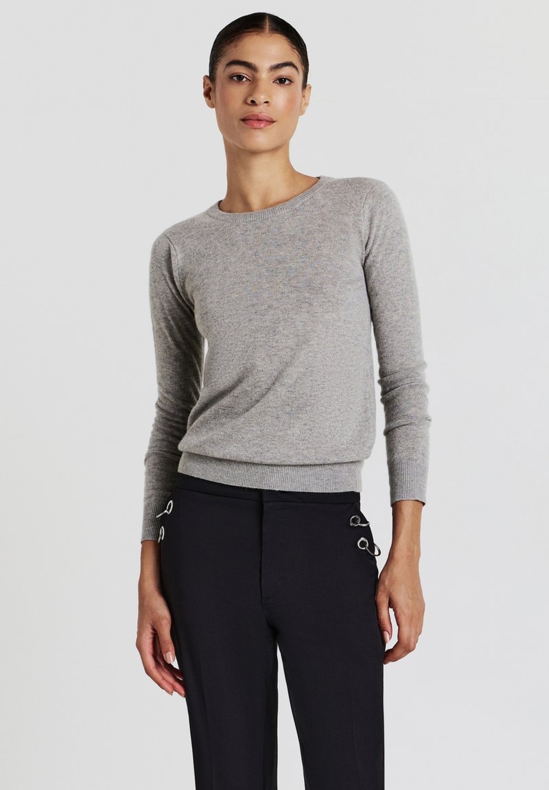 Maglione grigio aderente a maniche lunghe con scollo rotondo. Tessuto morbido e polsini a coste, abbinato a pantaloni neri su misura con dettagli in metallo argentato.