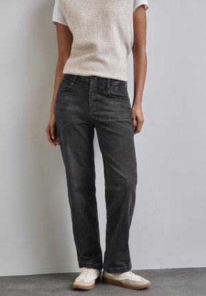 Person iført falmede sorte straight-leg jeans, hvide sneakers og en beige ærmeløs strik top, står ved en almindelig væg.