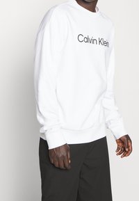 Witte sweatshirt van katoen, met een ronde hals, lange mouwen en een zwart "Calvin Klein" logo over de borst. Zachte textuur.