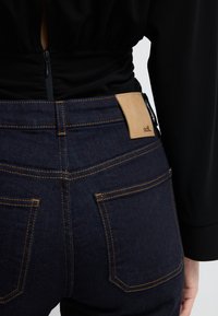 Tummansiniset farkut, joissa on kontrastiväri keltainen ommel. Ominaisuuksiin kuuluu vaaleanruskea nahkasaumapatch vyötäröllä ja kaksi takataskua.