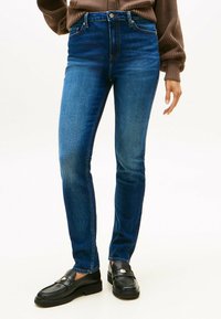 Jean en denim bleu avec une coupe ajustée, taille haute, design à cinq poches et décoloration subtile. Assorti à des mocassins noirs, mettant en valeur un look classique.