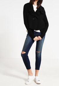 Chaqueta negra texturizada con detalles de volantes y un solo cierre, combinada con unos vaqueros ajustados oscuros rasgados y zapatillas blancas.
