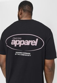 Svart bomulls T-skjorte med stort frontlogo i lys rosa som har "Club1990 apparel" og mindre tekst under som indikerer designets opprinnelse i Danmark.