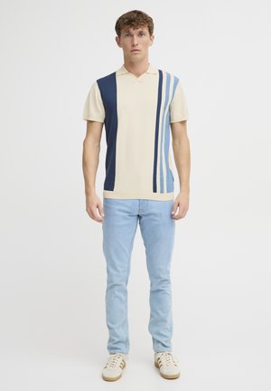 Gebreid poloshirt in crème met marineblauwe en blauwe verticale strepen. Korte mouwen en een zachte, gestructureerde afwerking. Gecombineerd met lichte jeans.