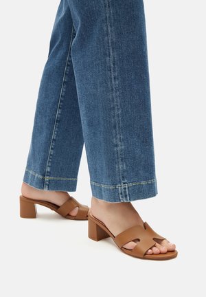 Piernas con jeans vaqueros azules y sandalias de tacón cuadrado color canela con tiras anchas cruzadas, de pie sobre una superficie blanca.