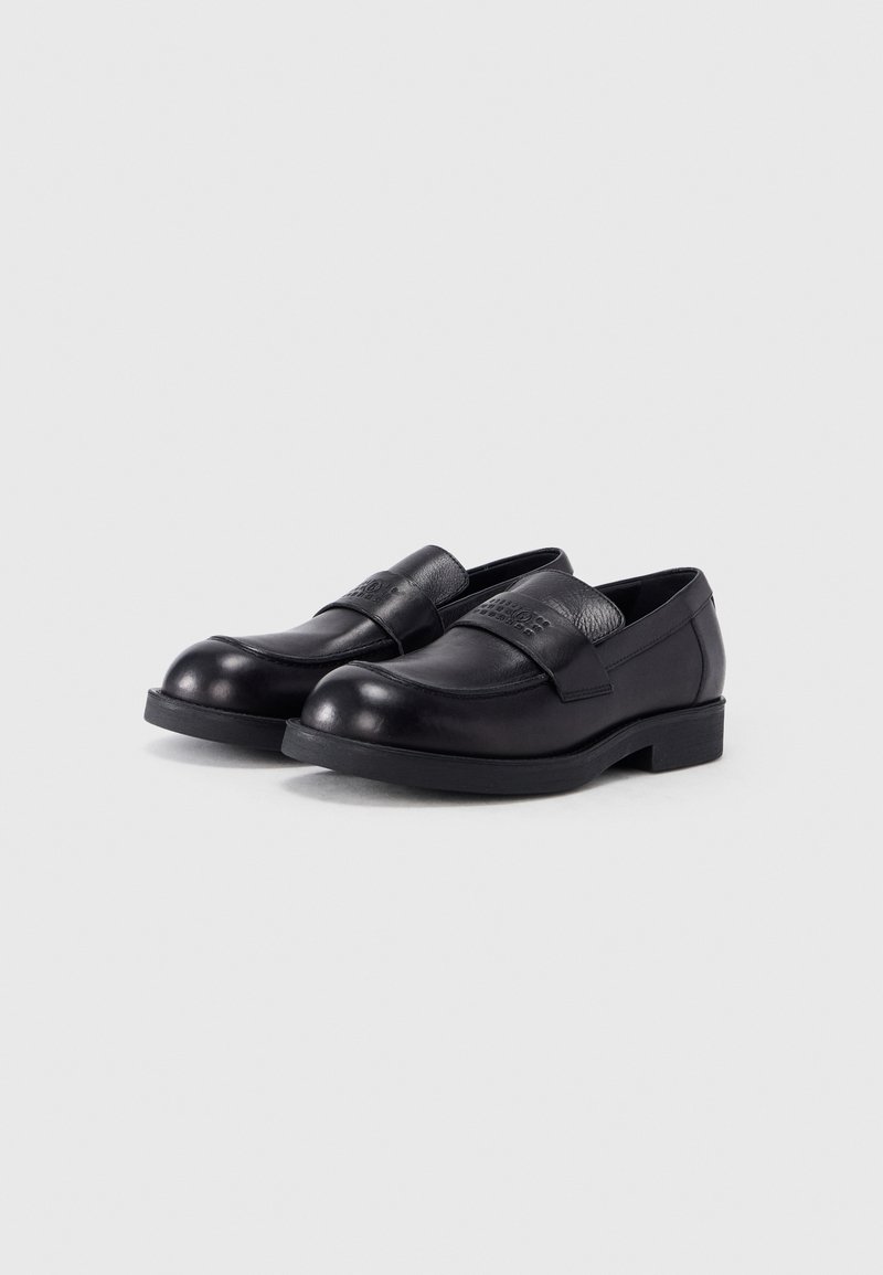 Par sorte læder slip-on loafers med rundet tå og en rem med præget mærke på en hvid baggrund.