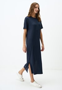 Abito blu navy, fino al ginocchio, realizzato in un morbido tessuto testurizzato con maniche corte e spacchi laterali. Indossato con sneakers bianche sportive.