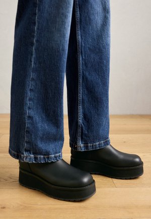 Füße mit schwarzen Plateauschuhen zum Hineinschlüpfen, kombiniert mit blauen weiten Jeans auf einem Holzboden.
