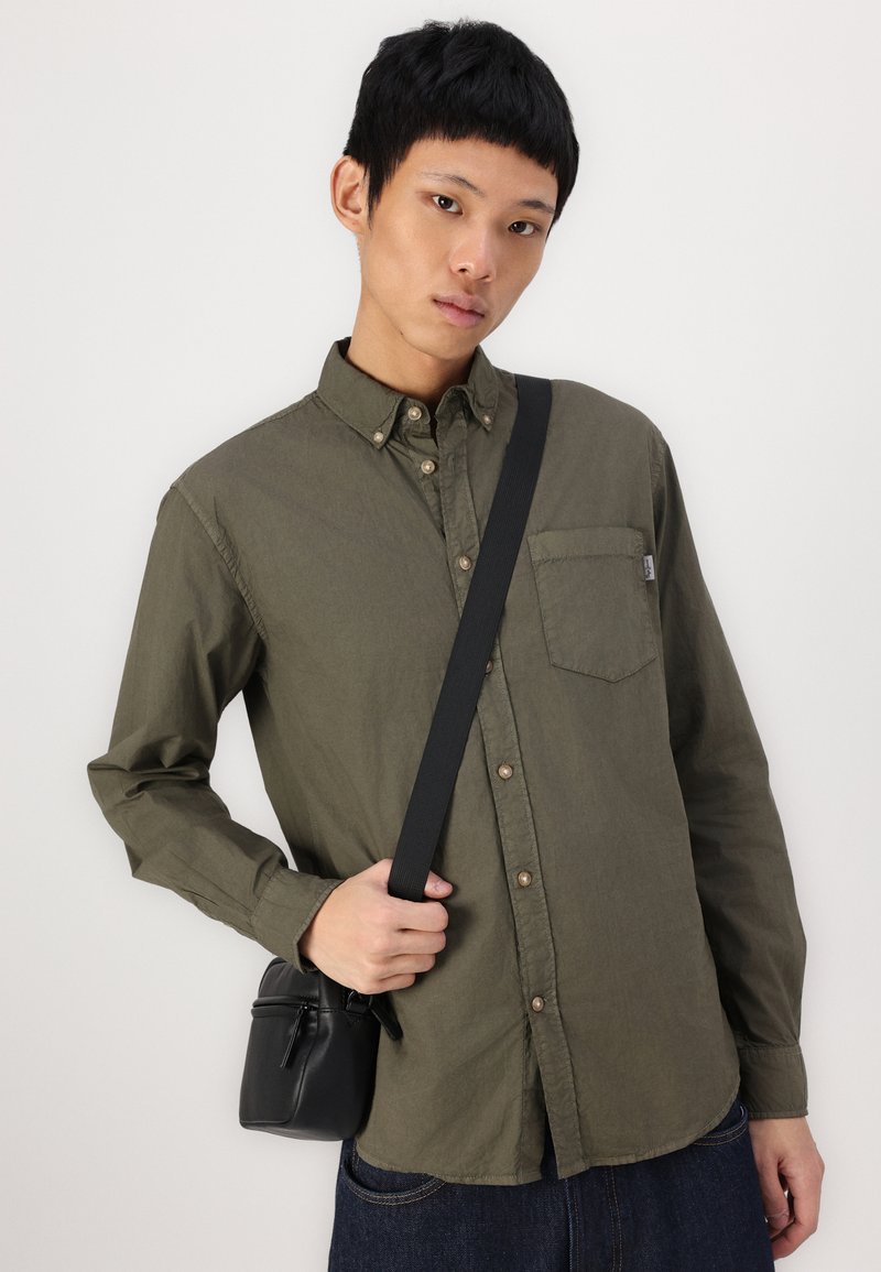 Olivgrüne Hemdbluse mit Brusttasche, kombiniert mit dunklen Jeans und einer kleinen schwarzen Umhängetasche mit Reißverschluss.