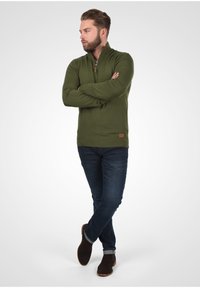 Olivgrüner Kapuzenpullover mit Reißverschluss, gerippten Bündchen und Saum, getragen über dunkelblauen Jeans und braunen Wildlederstiefeln, was eine lässige Silhouette zeigt.