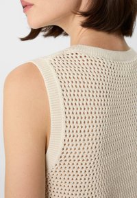 Knit sleeveless top in cream with a textured, open weave pattern, featuring a ribbed neckline and armholes.