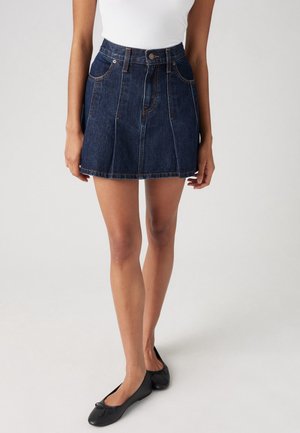 Femme portant une mini-jupe en jean bleu foncé, un haut blanc sans manches et des ballerines noires, debout sur un fond clair uni.