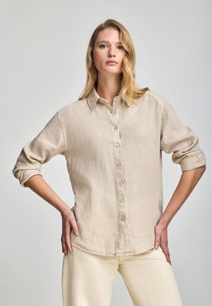 Beige Leinenhemd mit Knopfleiste und Kragen, langen Ärmeln, die bis zu den Ellenbogen gerollt sind, und einer subtilen Textur. Verfügt über ein kleines Logo-Detail nahe dem Saum.