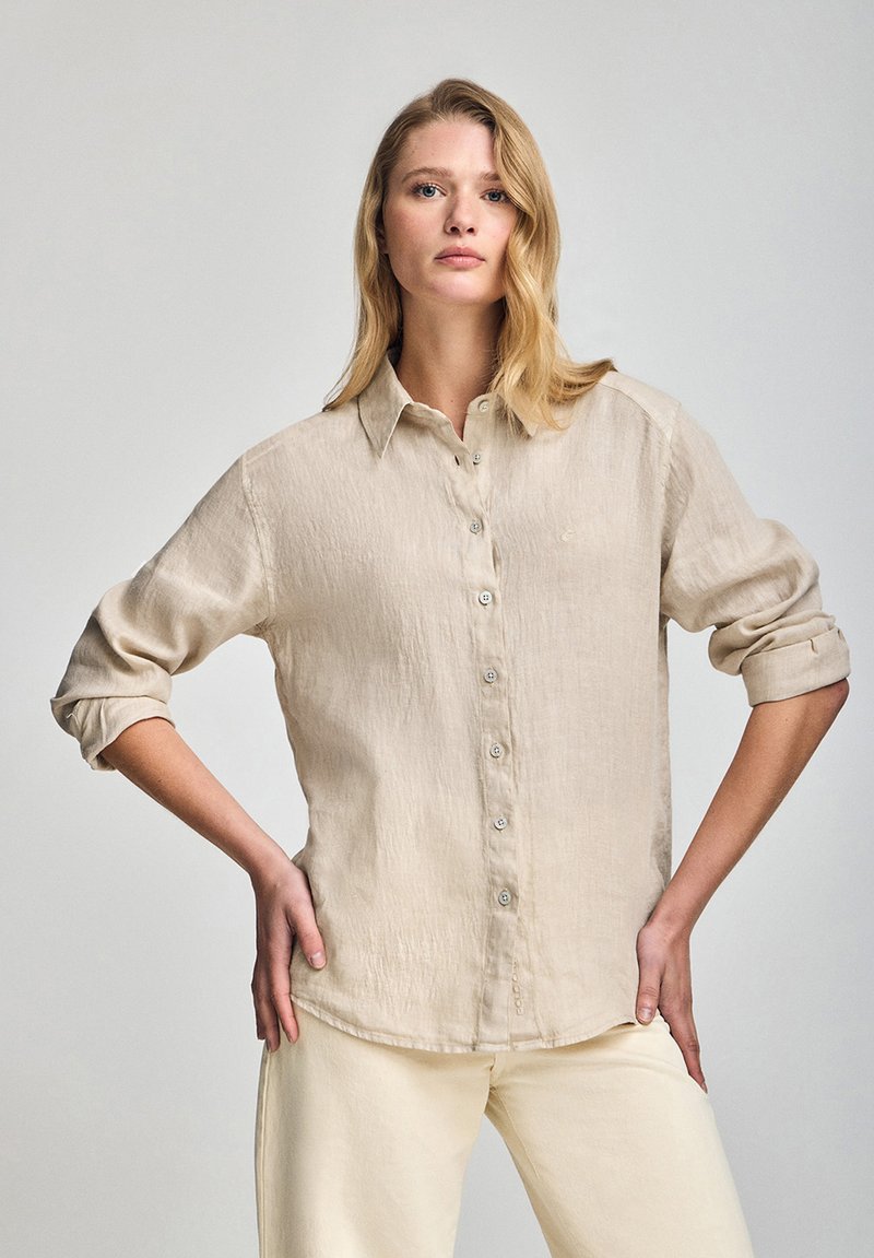 Camisa de lino beige con botones, cuello, mangas largas arremangadas hasta los codos y una textura sutil. Incluye un pequeño detalle de logo cerca del dobladillo.