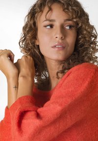 Pulover portocalie, pufoasă, cu mâneci largi. Modelul are păr texturat, creț și o mică tatuajă de cruce neagră pe încheietura mâinii. Machiaj natural.