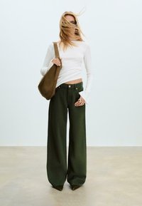 Žena s dlouhými blond vlasy, částečně zakrývajícími její obličej, má na sobě bílé tričko s dlouhým rukávem, volné tmavě zelené kalhoty, hnědou tašku a černé boty.