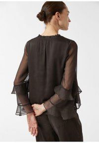 Zwarte, doorschijnende blouse met ruches aan de halslijn en wijde, gelaagde mouwen. Het materiaal lijkt licht en soepel, gedragen met donkere broek.