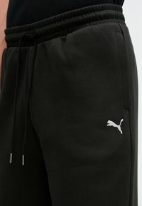 Czarne spodnie dresowe wykonane z miękkiego materiału, z regulowanym sznurkiem w talii, bocznymi kieszeniami oraz białym logo Puma wyszywanym na udzie.