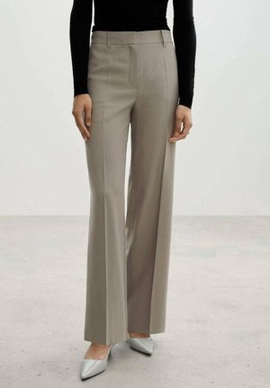 Pantalons beige ajustés avec un design évasé, texture lisse, plis sur le devant et taille haute, associés à des chaussures aiguës gris clair.