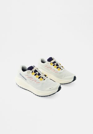 Par de zapatillas deportivas blancas con suelas negras, lazos amarillos, detalles en azul marino y la marca Salomon sobre un fondo blanco liso.