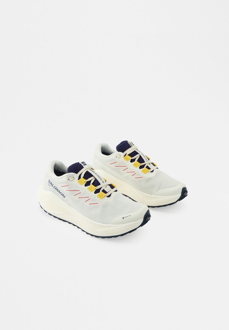 Valgeid jooksujalatseid mustade tallade, kollaste paeltõmmitsate, tumeroheliste detailide ja Salomoni logoga ühevärvilise valge taustal.