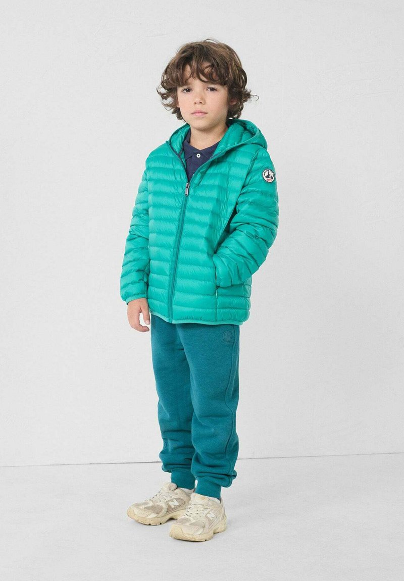 Turkooizen pufferjack met horizontale doorgestikte lijnen, ritssluiting aan de voorkant en capuchon. Gecombineerd met bijpassende turkooizen joggingbroek en beige sneakers.