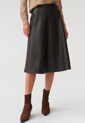 Jupe A-line en cuir noir avec une texture lisse, coupée au niveau des genoux, et une ceinture discrète. Associée à des bottines en daim marron à la cheville.