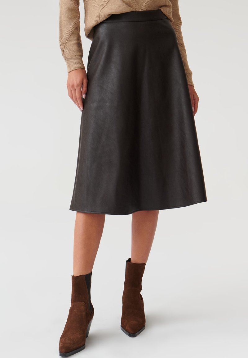 Jupe A-line en cuir noir avec une texture lisse, coupée au niveau des genoux, et une ceinture discrète. Associée à des bottines en daim marron à la cheville.