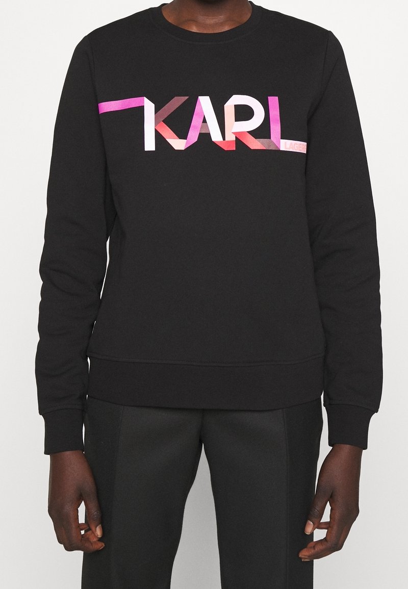 Sudadera negra con cuello redondo y puños de canalé, que presenta un colorido logo geométrico "KARL" en el pecho en tonos rosa, rojo y púrpura.