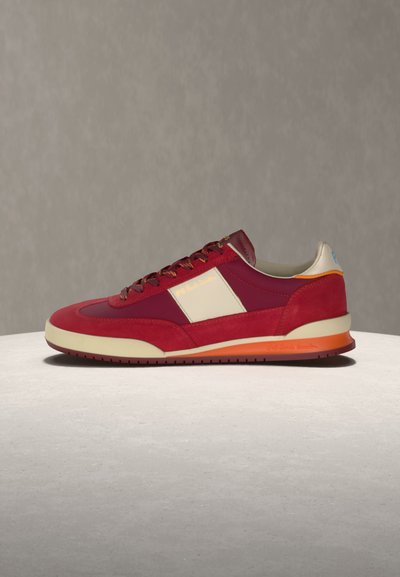 Κόκκινο και βουργουνδί αθλητικό παπούτσι με κρεμ πλαϊνή επιφάνεια και πορτοκαλί λεπτομέρεια στη σόλα, τοποθετημένο σε ανοιχτό γκρι επιφάνεια πάνω σε ουδέτερο φόντο.