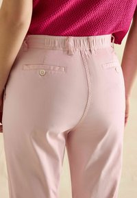 Gaiši rozā bikses ir ar elastīgu jostu, divām aizmugurējām kabatām ar pogu aizdari un gludu, nedaudz spīdīgu auduma tekstūru.