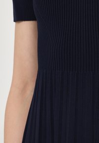 Rippengesprenkeltes marineblaues Kleid mit kurzen Ärmeln, einem figurbetonten Oberteil und einem ausgestellten Rock mit vertikalen Falten. Hergestellt aus einem glatten, strukturierten Stoff.