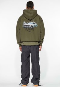 Olivengrønn hettegenser med grafikk av en vintagebil på baksiden, sammen med mørke cargo-bukser og sneakers med oransje såler.