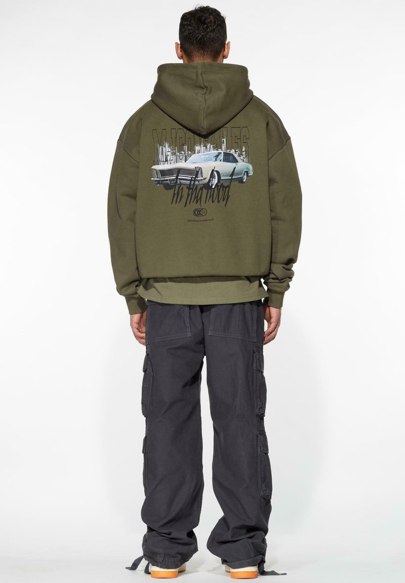 Olivgrön hoodie med en vintage bilgrafik på baksidan, kombinerad med mörka cargo-byxor och sneakers med orange sål.