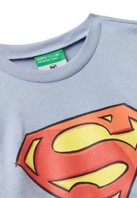 Jasnoniebieska koszulka z czerwoną i żó�łtą grafiką logo Supermana z przodu oraz zieloną metką United Colors of Benetton wewnątrz kołnierza.