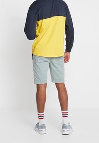 Camisa de manga larga en bloques de color, con la parte superior en navy y la inferior en amarillo, combinada con pantalones cortos verdes claros y zapatillas grises. Calcetines a rayas rojas.