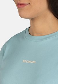 Camiseta de algodón azul clara con cuello redondo y mangas cortas, que presenta el nombre de la marca "ROSSIGNOL" en texto blanco en la parte frontal.