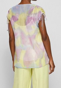 Blouse transparente dans un motif tie-dye pastel jaune, rose et bleu, avec des détails à cordon au niveau des manches et une coupe décontractée.