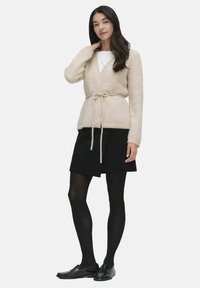Kremowy kardigan z paskiem, o strukturalnym splocie, z dekoltem w serek i długimi rękawami, w połączeniu z czarną spódnicą i czarnymi rajstopami.