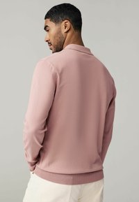 Lyserød poloshirt med lange ærmer, tekstureret stof, ribbede manchetter og kant samt en klassisk krave. Vist fra en bagvinkel.