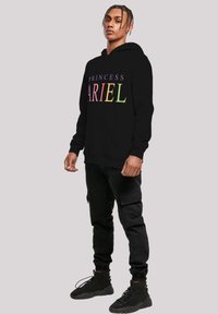 Čierna mikina má na sebe text "PRINCEZNA ARIEL" v pestrofarebných písmenách, v kombinácii s čiernymi cargo nohavicami a čiernymi vysokými teniskami.