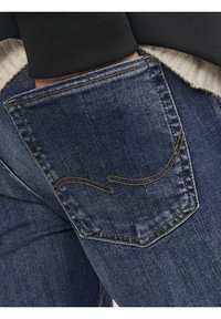 Jeans de mezclilla azul oscuro que presentan un bolsillo trasero con un patrón de costura curvado en hilo naranja contrastante, destacando el diseño y la textura del bolsillo.