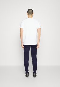 Weißes Baumwoll-T-Shirt kombiniert mit navyblauen, taillierten Hosen, die beide ein einfaches Design aufweisen. Die Schuhe haben schwarze Akzente und ein Logo.
