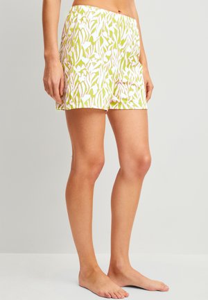 Frau, die grün-weiß gemusterte Shorts mit Blumen trägt, steht barfuß vor einem schlichten hellgrauen Hintergrund.
