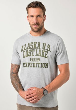 Mand med skæg og tatoveret venstre arm, iført grå t-shirt med teksten "Alaska U.S. Lost Lake Trail Expedition" og et armbåndsur, står med hænderne foldet.