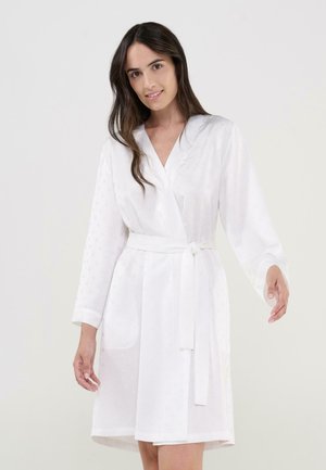 Mujer con el cabello largo y oscuro, vistiendo una bata blanca de manga larga con cinturón, de pie contra un fondo claro y liso.