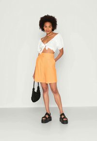 Top branco cropped com laço na frente, calções laranja de cintura alta, sandálias pretas chunky e uma mala de ombro preta texturizada. Fundo neutro.