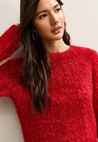 Czerwony, puszysty sweter wykonany z teksturowanej, miękkiej tkaniny z klasycznym okrągłym dekoltem. Luźny krój i długie rękawy podkreślają przytulny charakter ubrania.